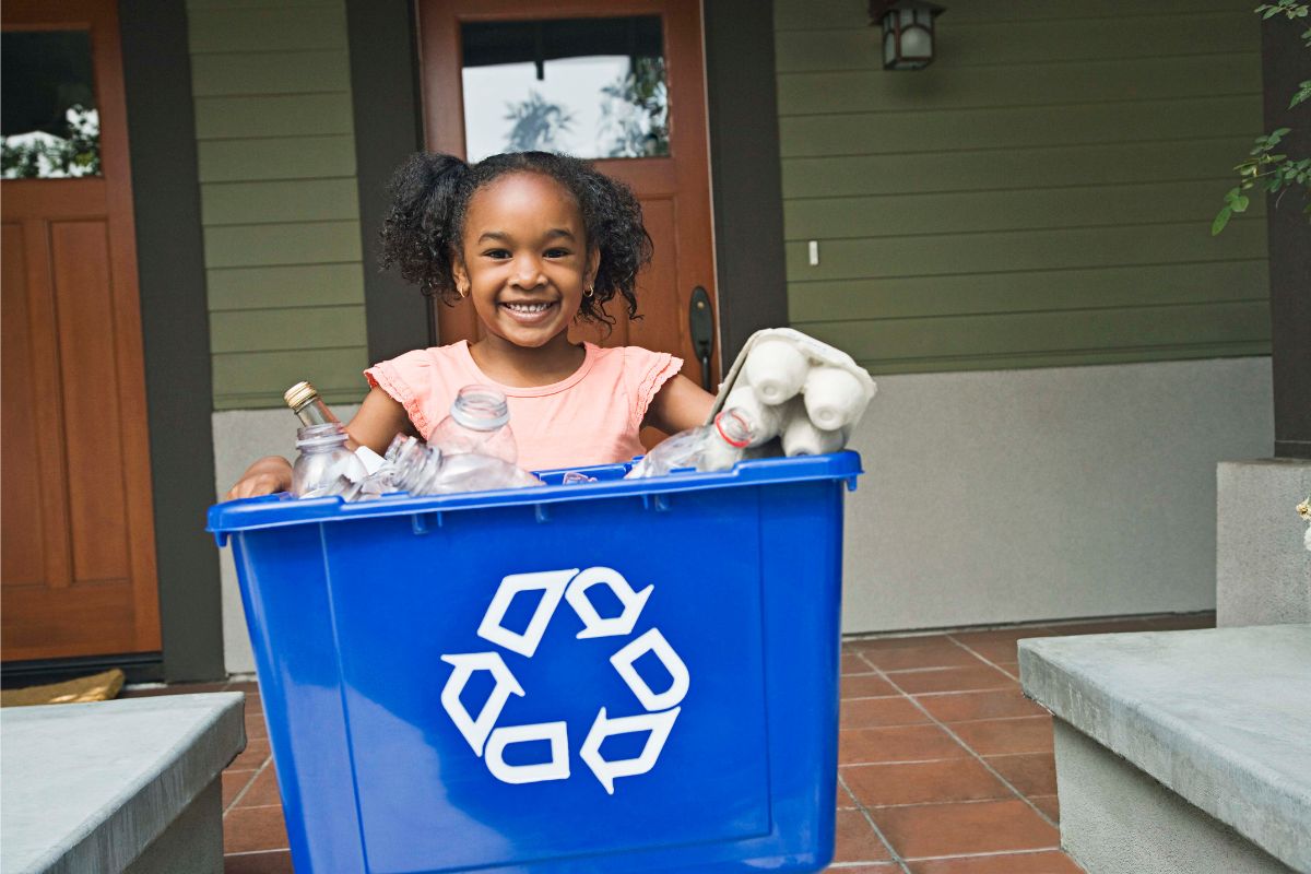 A smiling child holds a blue recycling bin filled with glass and plastic bottles and a carton outside a house with a green exterior and wooden door, embracing sustainable living practices.