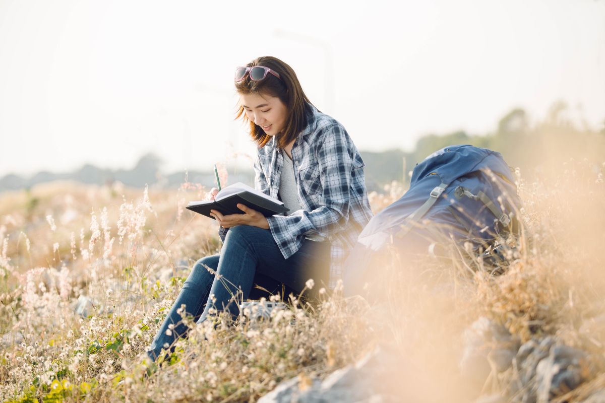 A person with a backpack sits outdoors on a grassy field, writing eco friendly travel tips in a notebook. They are wearing a plaid shirt and sunglasses on their head. The background features a hazy sky and distant trees.