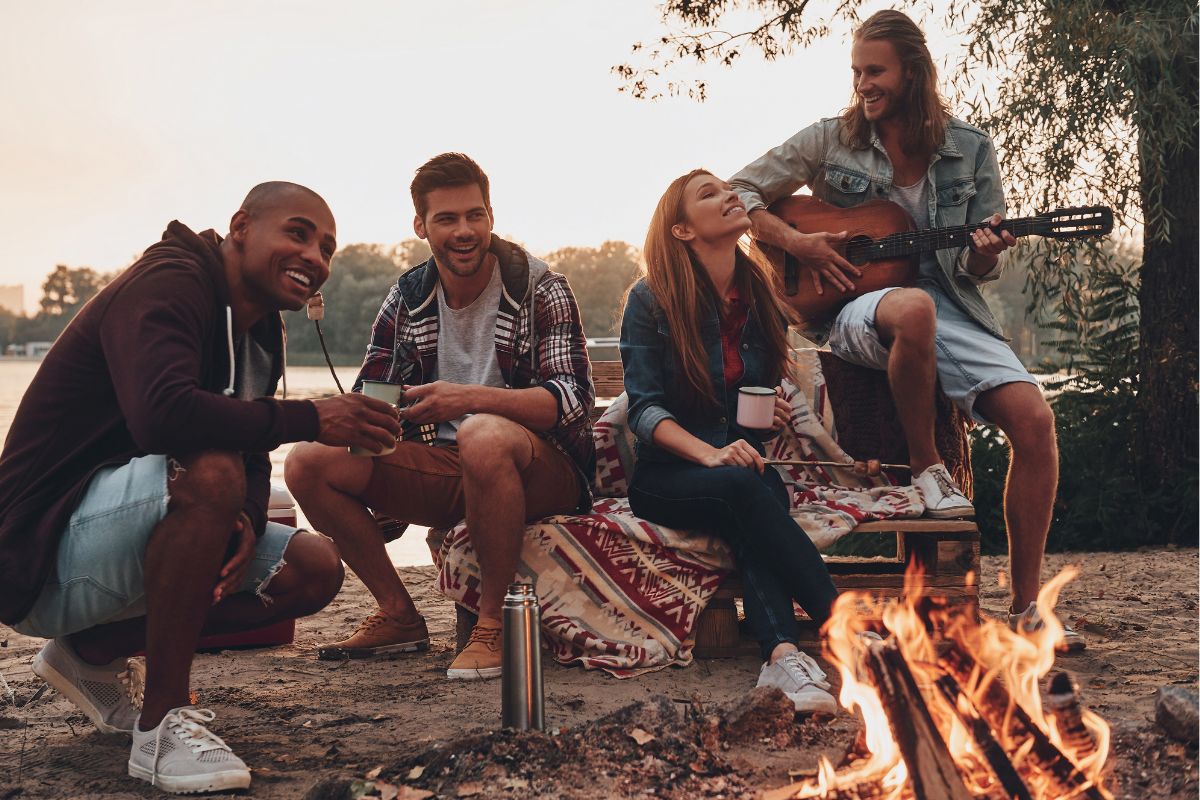 Four people sitting around a campfire, one playing a guitar while others smile and chat, enjoying activities in the Great Smoky Mountains, surrounded by nature.