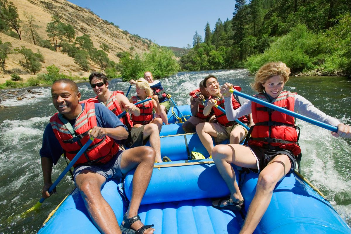 A group of six people wearing life jackets are white-water rafting on a river surrounded by trees and hills. They are paddling with blue oars and appear to be enjoying the experience, one of many thrilling activities in the Great Smoky Mountains.