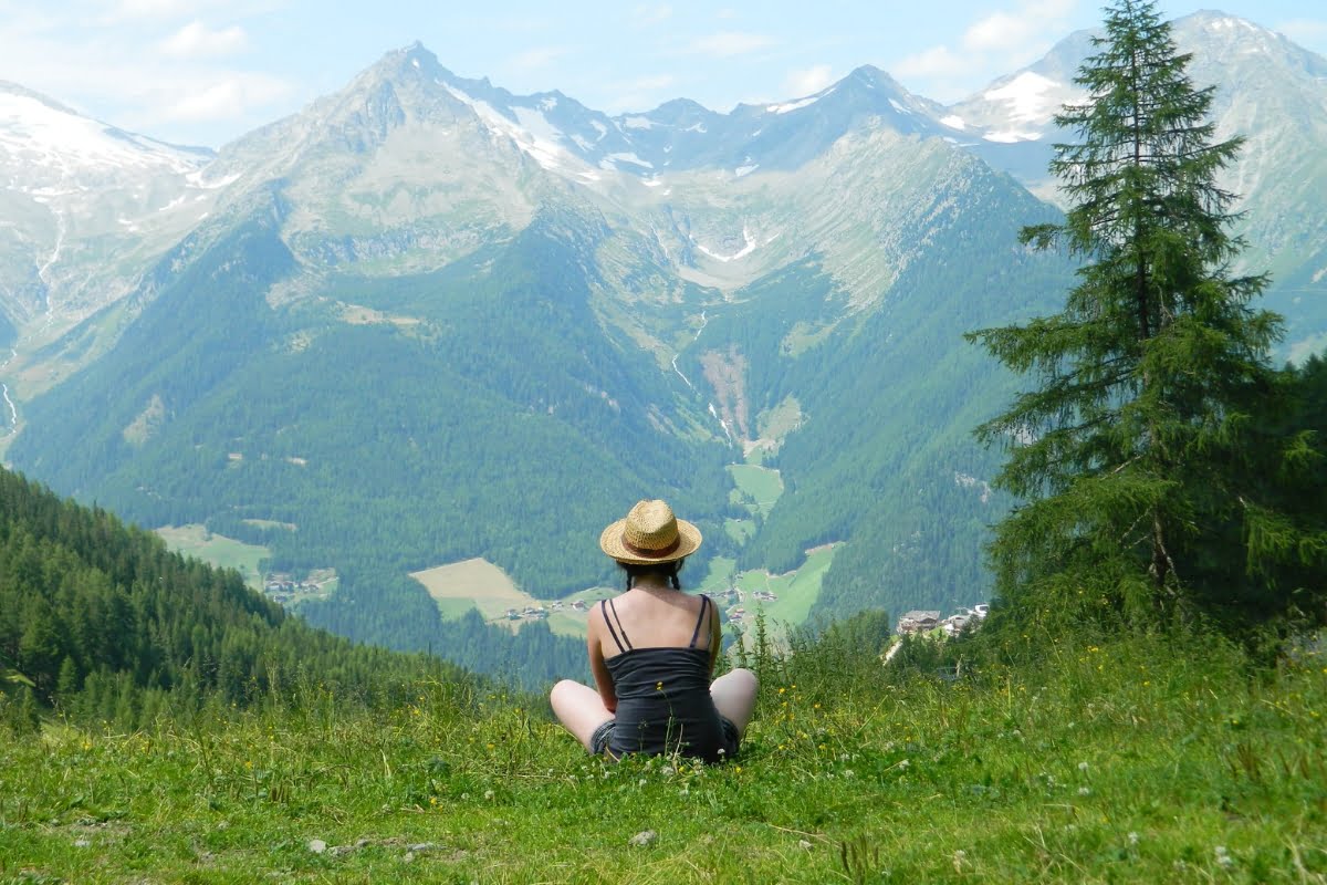 Person wearing a straw hat and black clothing sits on a grassy hillside, overlooking a valley with scattered houses and surrounded by large, green mountains, contemplating the region's wildlife conservation efforts.