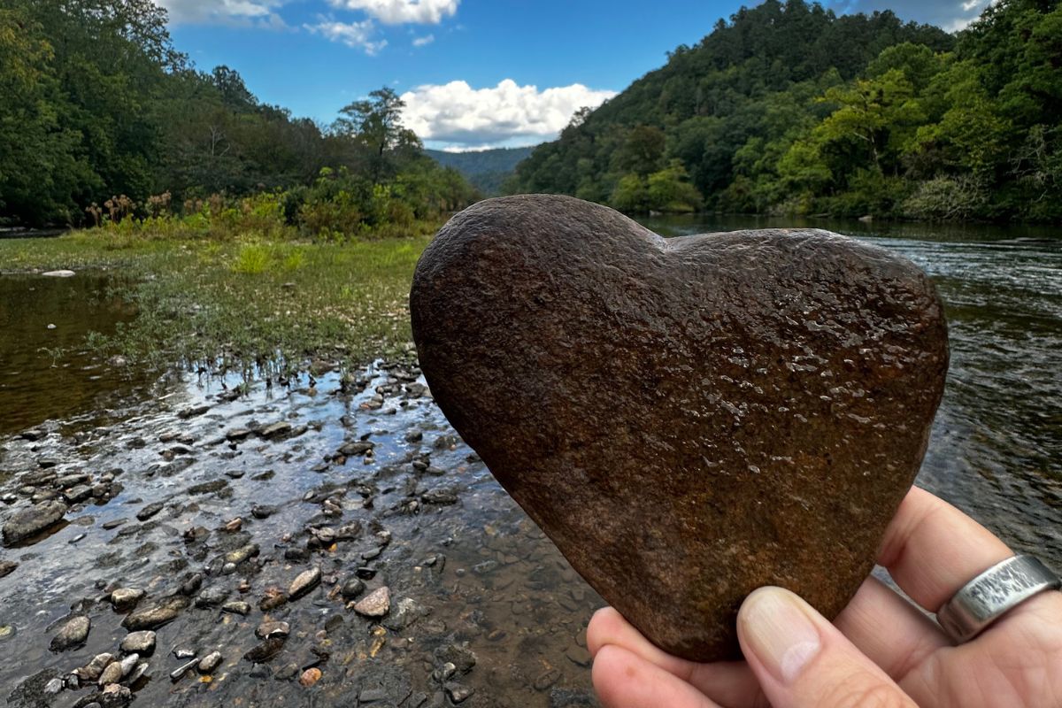 A hand holding a heart-shaped rock near a river with forested hills and a cloudy sky in the background, embodying the essence of natural health and nature experiences in Basecamp Smoky Mountains.