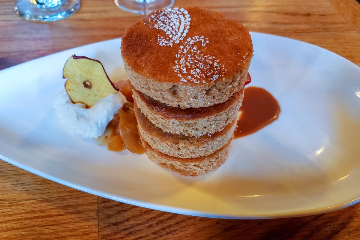 A dessert plate featuring a stack of three round cakes, dusted with powdered sugar, alongside a dollop of cream, an apple chip, and some caramel sauce offers delightful culinary experiences.