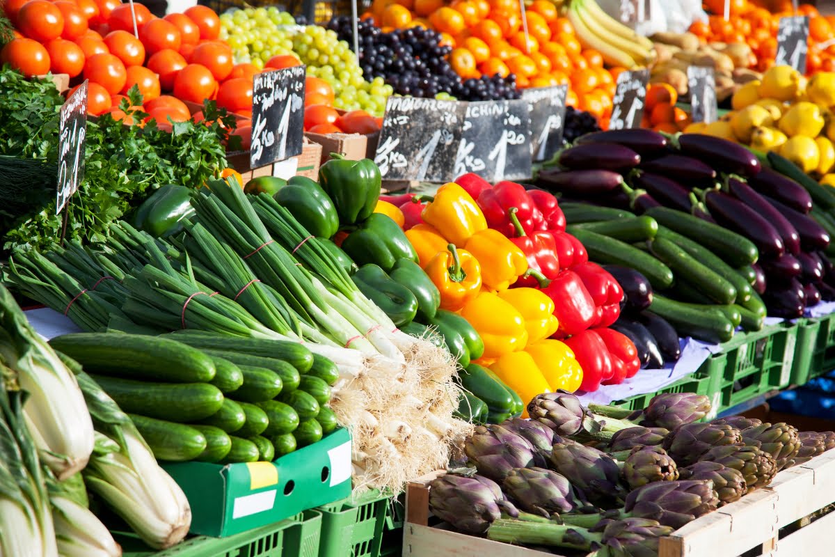 A colorful display of fresh vegetables and fruits at a market stall, including green onions, bell peppers, eggplants, zucchini, tomatoes, herbs, and various fruits, invites delightful culinary experiences.