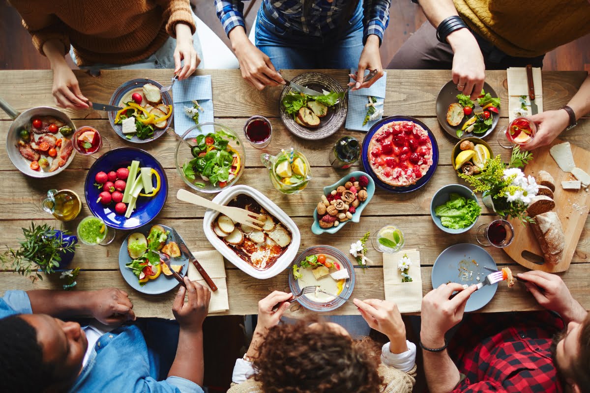 A group of people sit around a wooden table filled with various dishes, including salads, bread, fruits, baked goods, and drinks, engaging in rich culinary experiences.