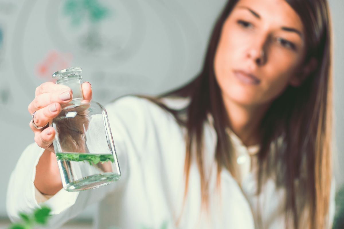 A woman in a lab coat examines a glass container with a liquid and plant inside, promoting natural health against a lab background.