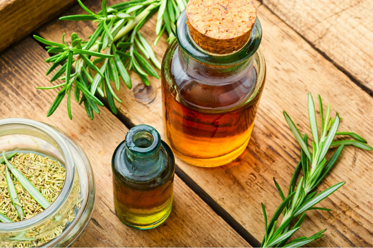 Two corked glass bottles filled with herbal oil and a small jar containing dried herbs rest on a wooden surface, promoting natural health. Fresh rosemary sprigs are artfully arranged around them.