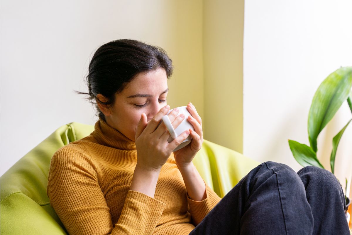 A person wearing a mustard sweater sits on a green chair, sipping from a white mug, with a small plant visible in the background, embodying the essence of natural health.