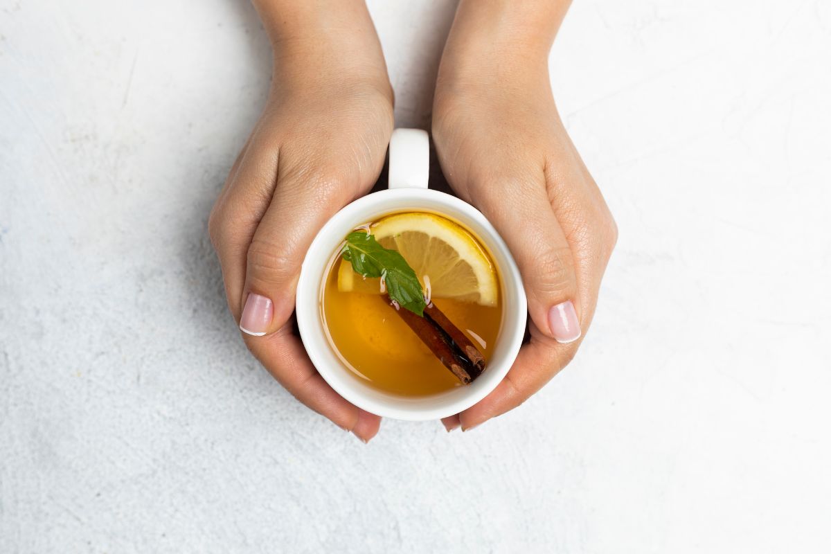 Two hands holding a white mug filled with tea, garnished with a lemon slice, a cinnamon stick, and a fresh mint leaf on a light background—an inviting scene that embodies natural health.