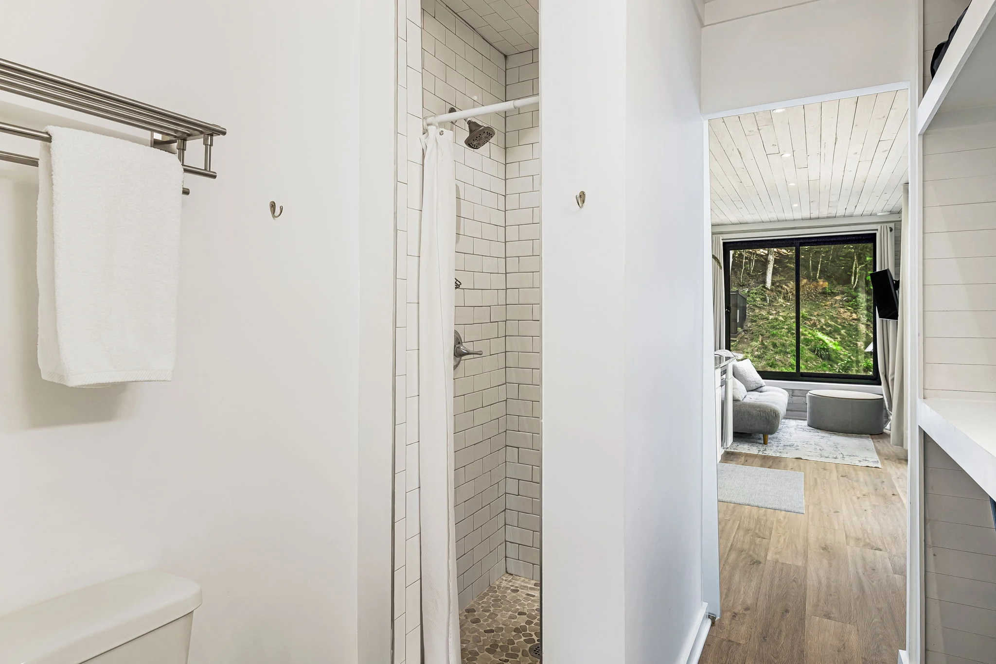 A small bathroom with a white tile shower, towel rack holding a white towel, and an adjacent living area with large windows and forest view. Wood-grain floor extends from bathroom to living area.