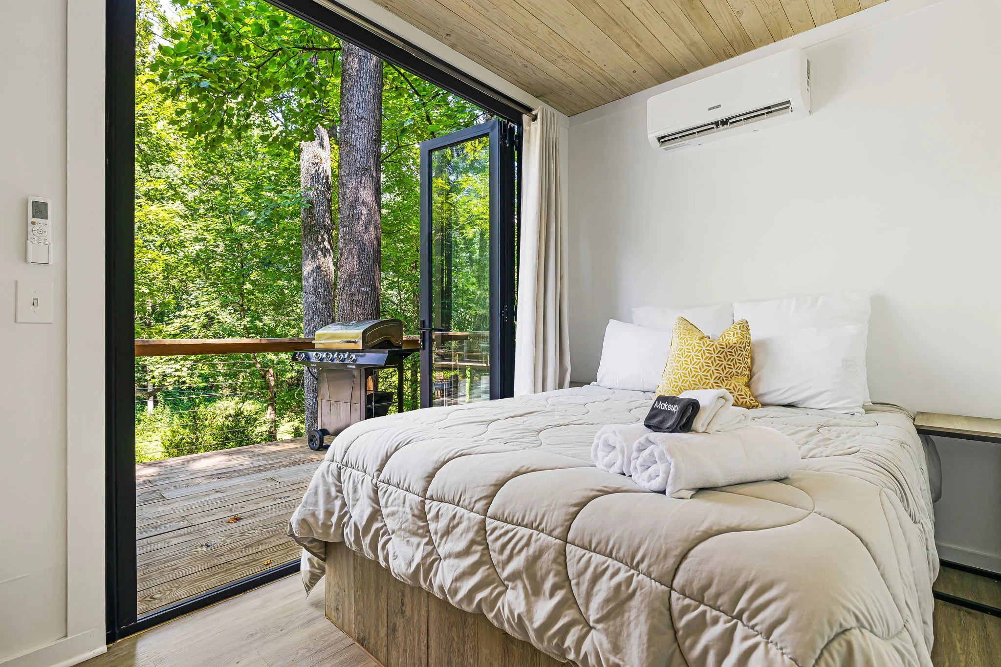 A cozy bedroom with a neatly made bed, adjacent to an open sliding glass door revealing a wooden deck with a barbecue grill and lush green trees outside.