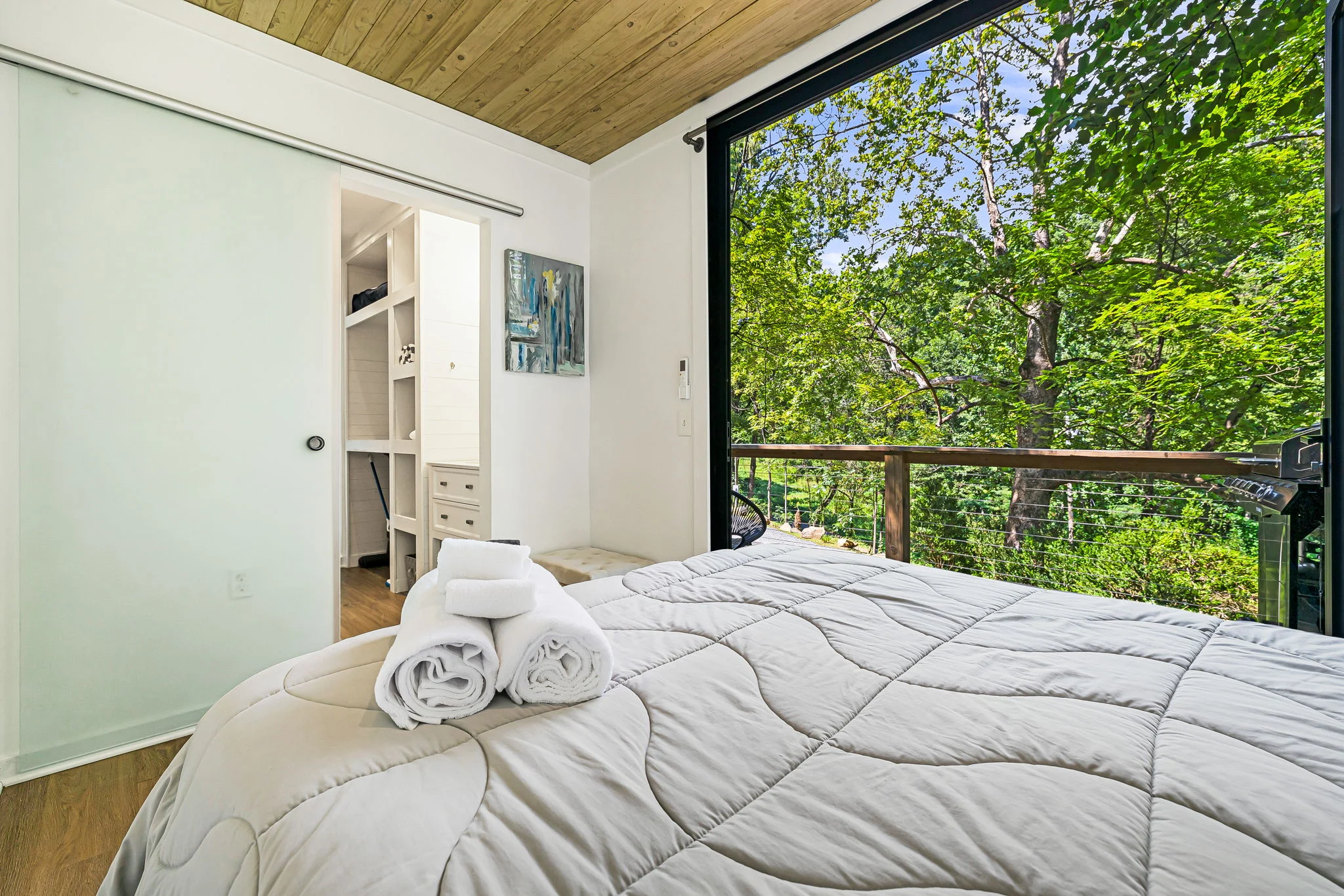 A bedroom with a large window overlooking trees, featuring a bed with white linens and towels, and a sliding door leading to a closet.