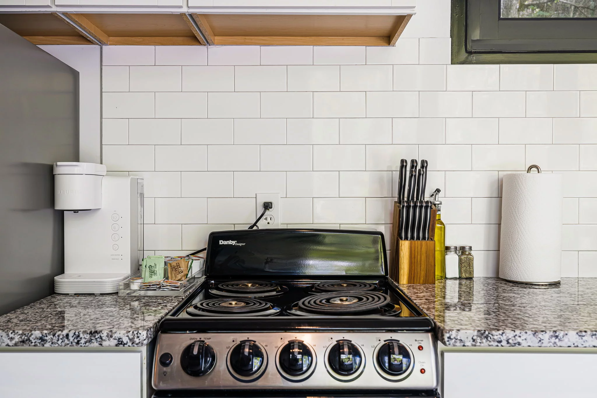 A kitchen counter in an eco-villa features a coffee maker, spice jars, knife block, paper towel holder, and a black electric stove with four burners set against a white subway tile backsplash for an elevated cooking experience.
