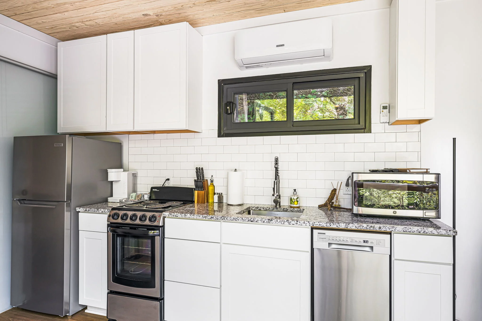 Modern kitchen with white cabinets, a stainless steel fridge, stove, dishwasher, and a countertop with kitchen utensils. A window is above the sink, providing an elevated view that complements this eco-villa's sustainable design. An air conditioning unit sits above the window for added comfort.