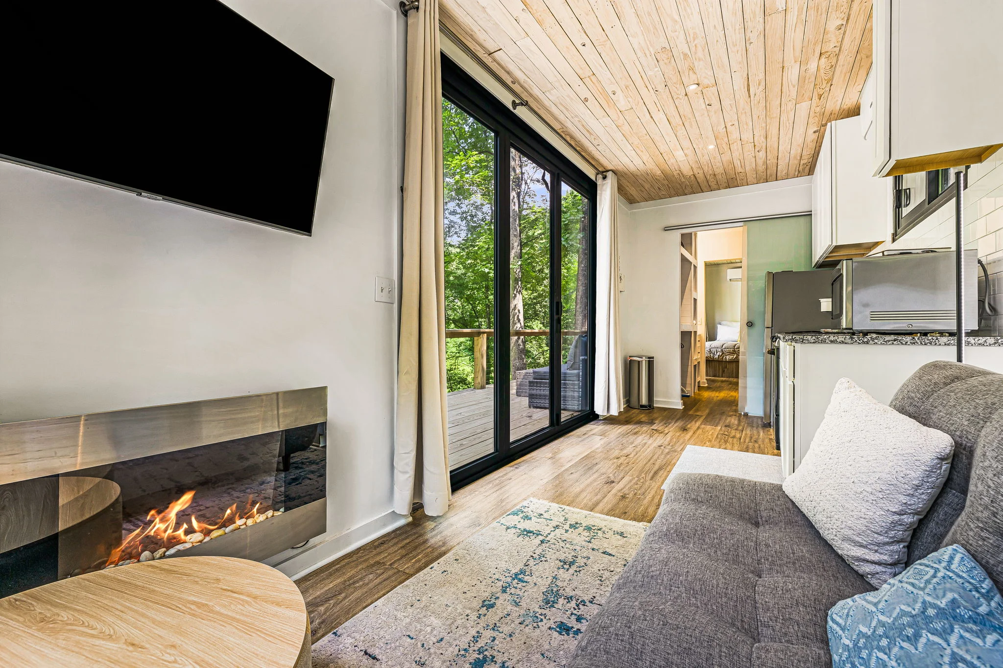 Compact living room with a sofa, wall-mounted TV, and fireplace, leading to a balcony with forest view through large glass doors. Small kitchen area visible on the right. Light wood ceiling and floor.