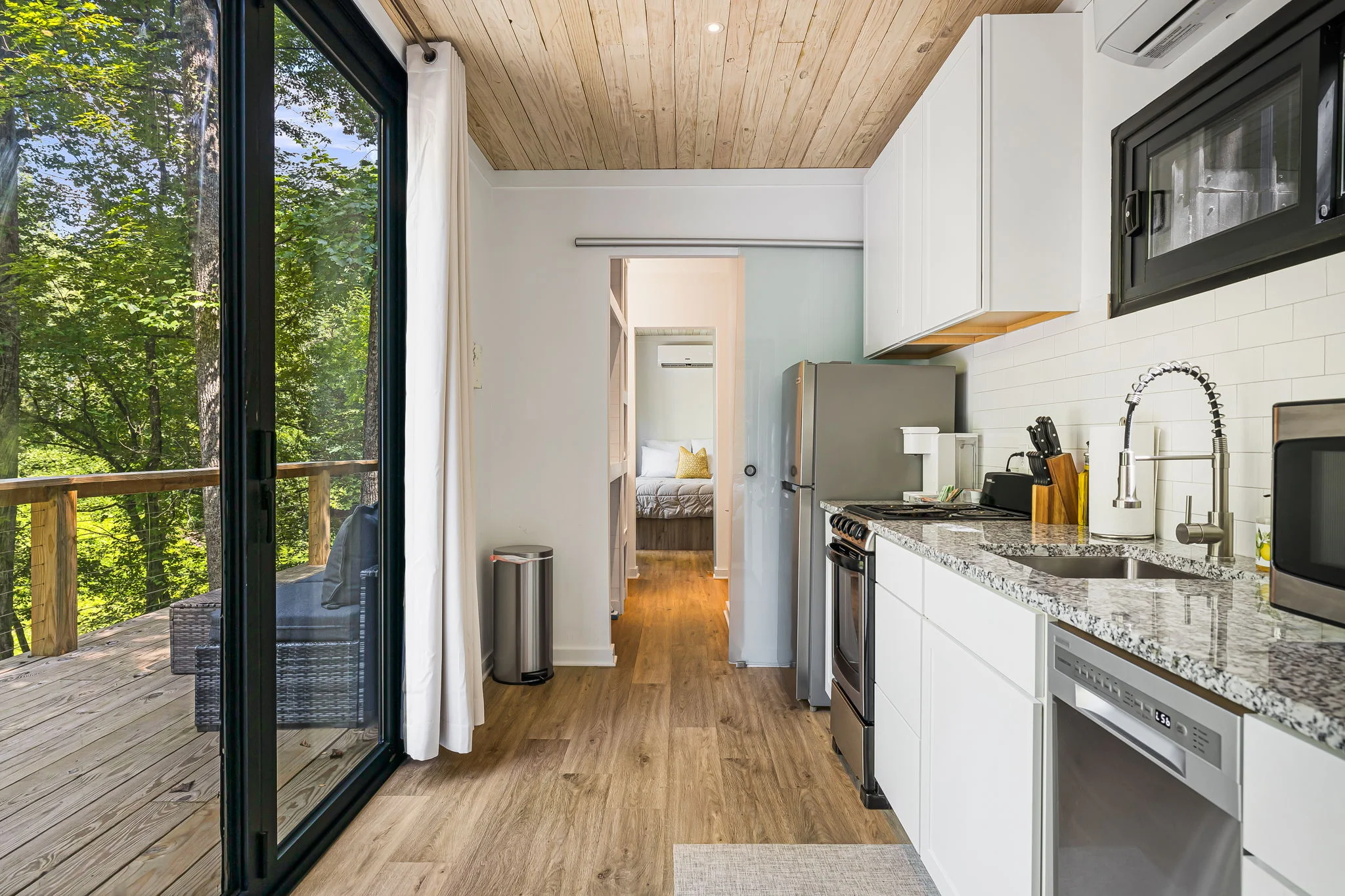 A small modern kitchen with stainless steel appliances, white cabinetry, and a stone countertop. A sliding glass door leads to a wooden deck with outdoor seating and a forest view. A bedroom is visible in the background.