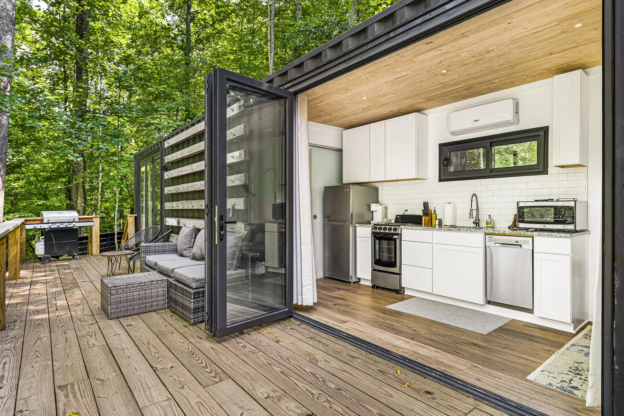 An open small house with a modern kitchen and a sitting area on a wooden deck surrounded by trees.