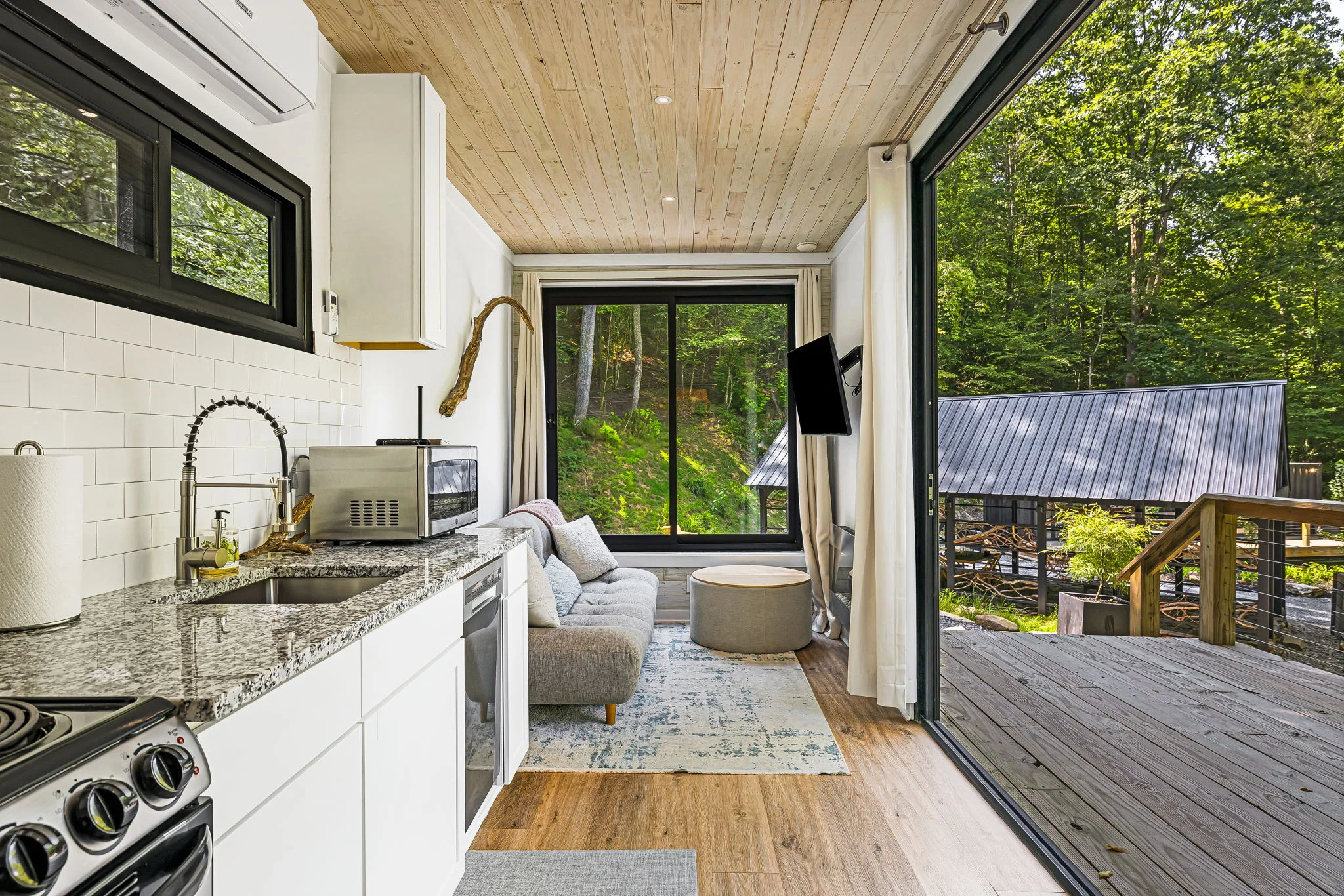 Modern tiny house interior featuring a small kitchen with granite countertops, stove, and microwave; adjacent to a cozy seating area with large sliding glass doors opening to a wooded deck area.