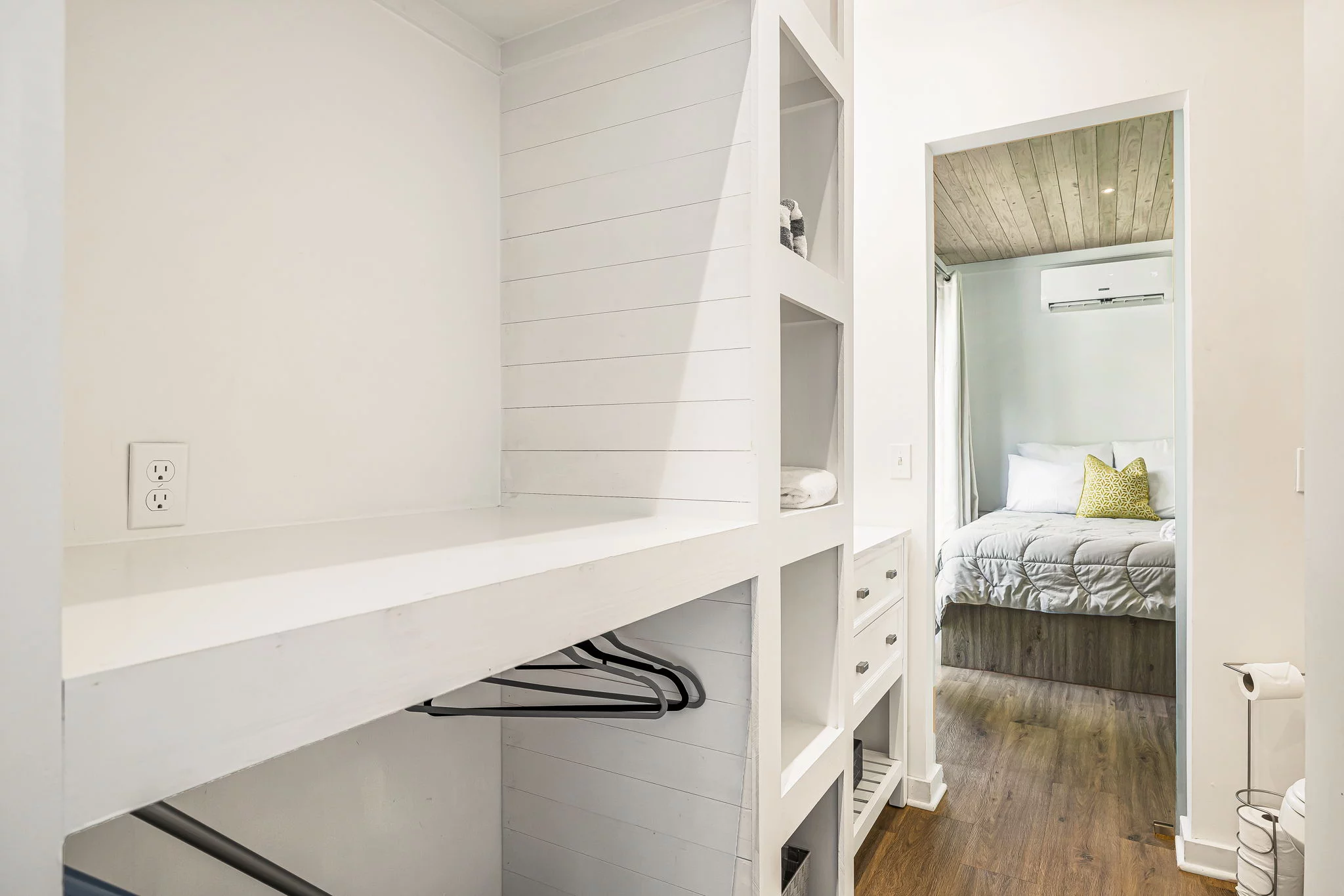 Bright walk-in closet with built-in shelves, hanging space, and an adjacent view of a rustic bedroom featuring a wooden ceiling and bed.