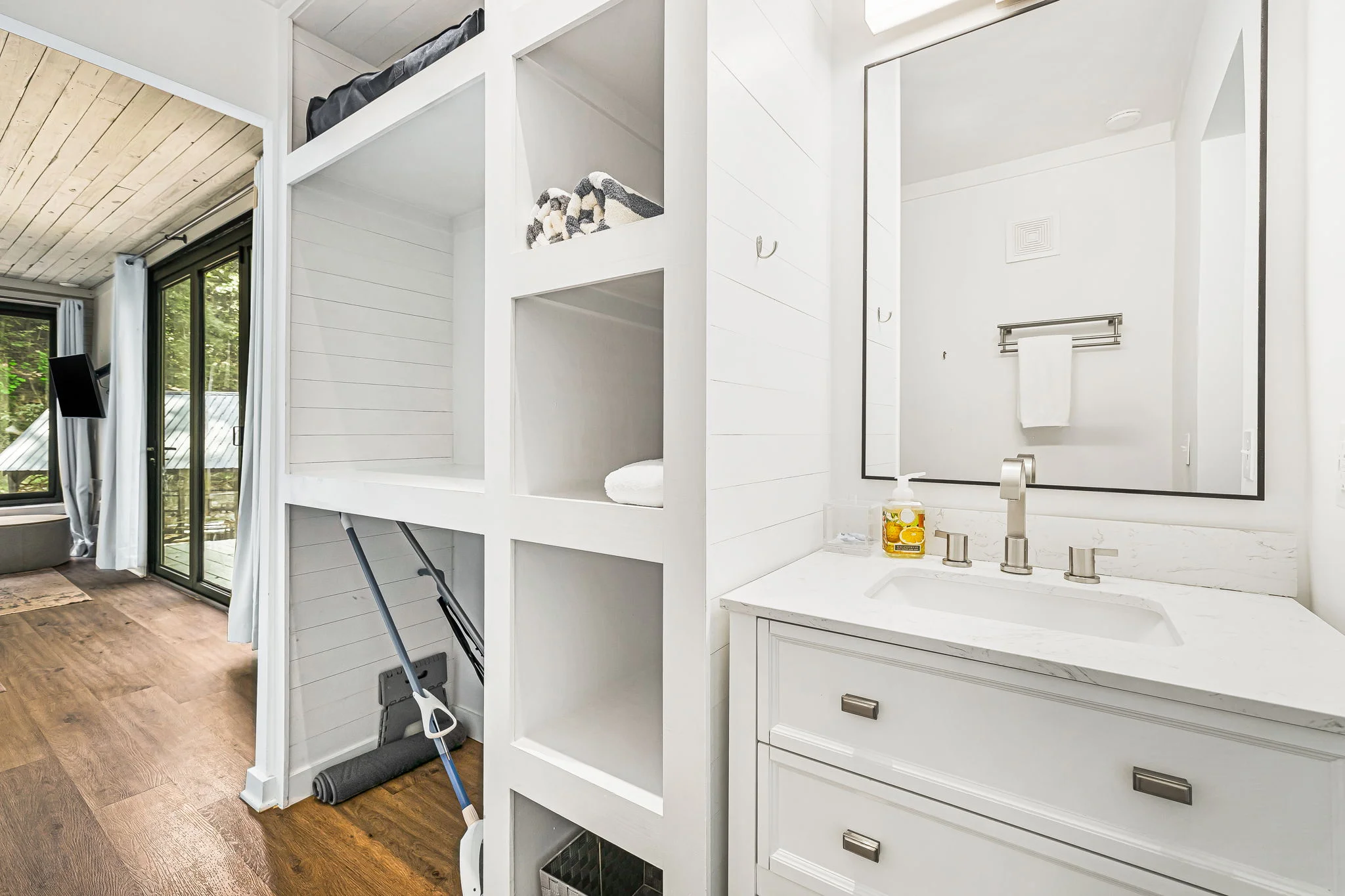 A tidy bathroom features a white vanity with a sink and faucet, a mirror above, and shelves for storage. To the left, an open doorway leads to a living area with large windows.