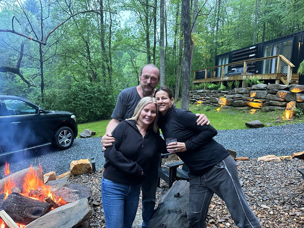 Three people stand together outdoors near a campfire, with greenery and a modern cabin in the background. Dressed in casual clothes, they appear to be enjoying their retreat setting.