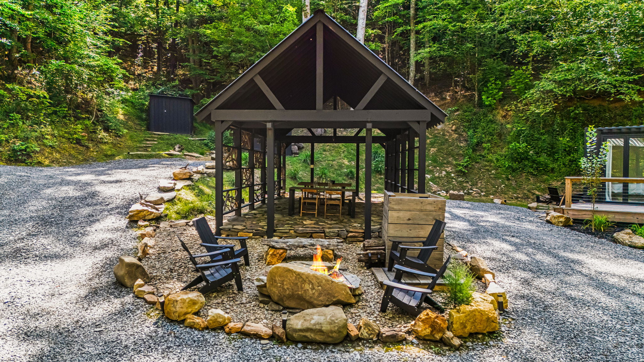 A small wooden gazebo with outdoor seating and a fire pit is surrounded by gravel paths in a wooded area. Two black chairs face the fire pit, and there's a small wooden structure in the background.