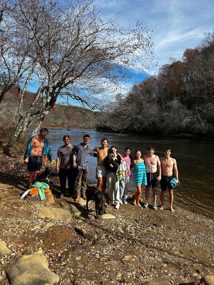 A group of nine people and a dog stand by a riverbank with mostly bare trees in the background. Some individuals wear swimsuits, while others are clothed, suggesting a mix of activities often seen in retreats near the water.