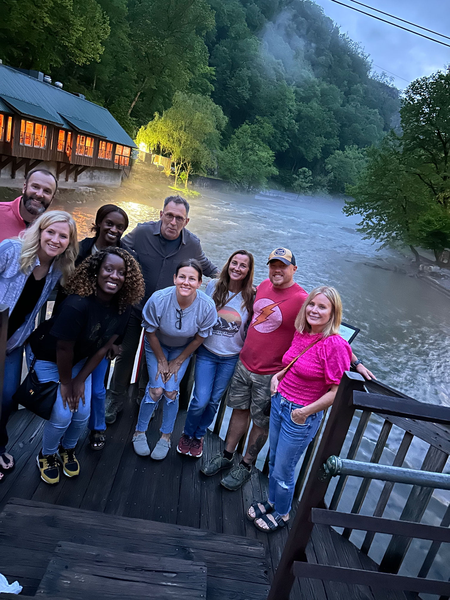 A group of nine people stands on a wooden deck by a river, enjoying the serenity typical of retreats, with a building and lush trees in the background.