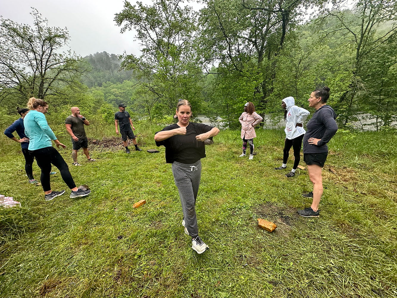 A group of people standing in a grassy outdoor area surrounded by trees, engaging in a fitness activity. One person is in the foreground with arms raised, while others stand and observe, exemplifying the spirit of wellness retreats and group activities.