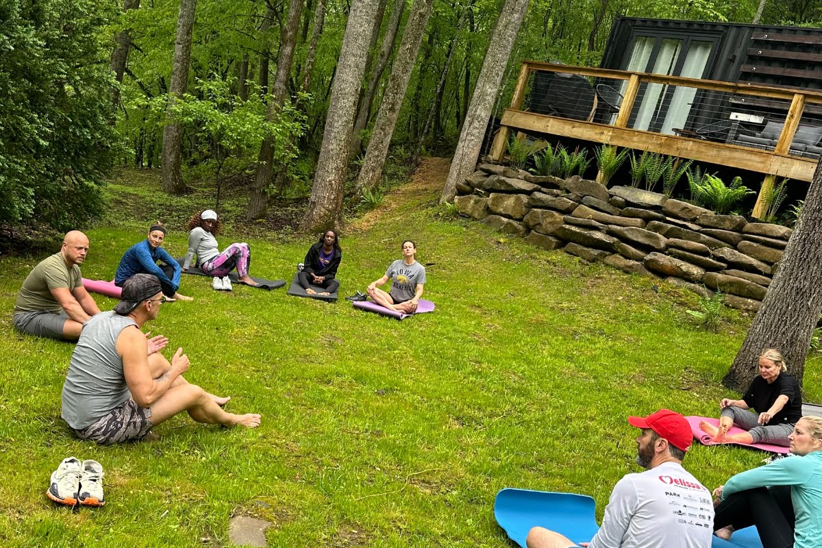 A group of people sitting in a circle on yoga mats in a grassy area near a cabin in the woods, enjoying one of their serene retreats.