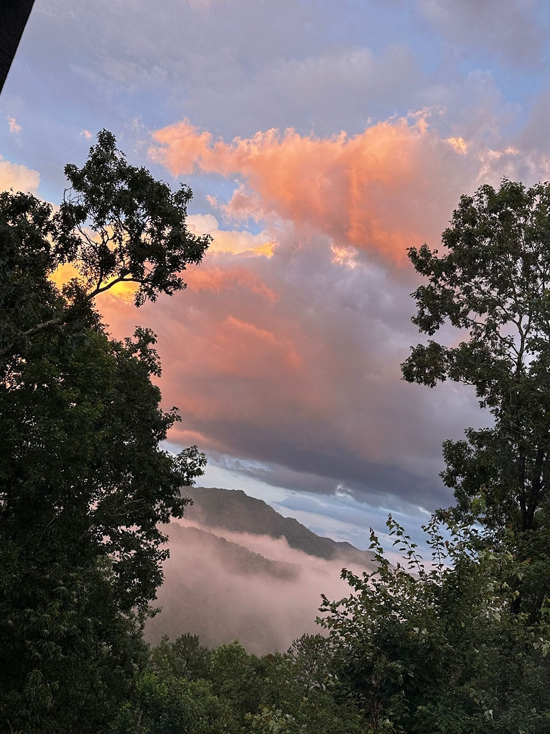 View of a misty mountain landscape at sunset, with vibrant orange clouds and trees framing the scene—perfect for a peaceful nature retreat in the heart of the Smoky Mountains.