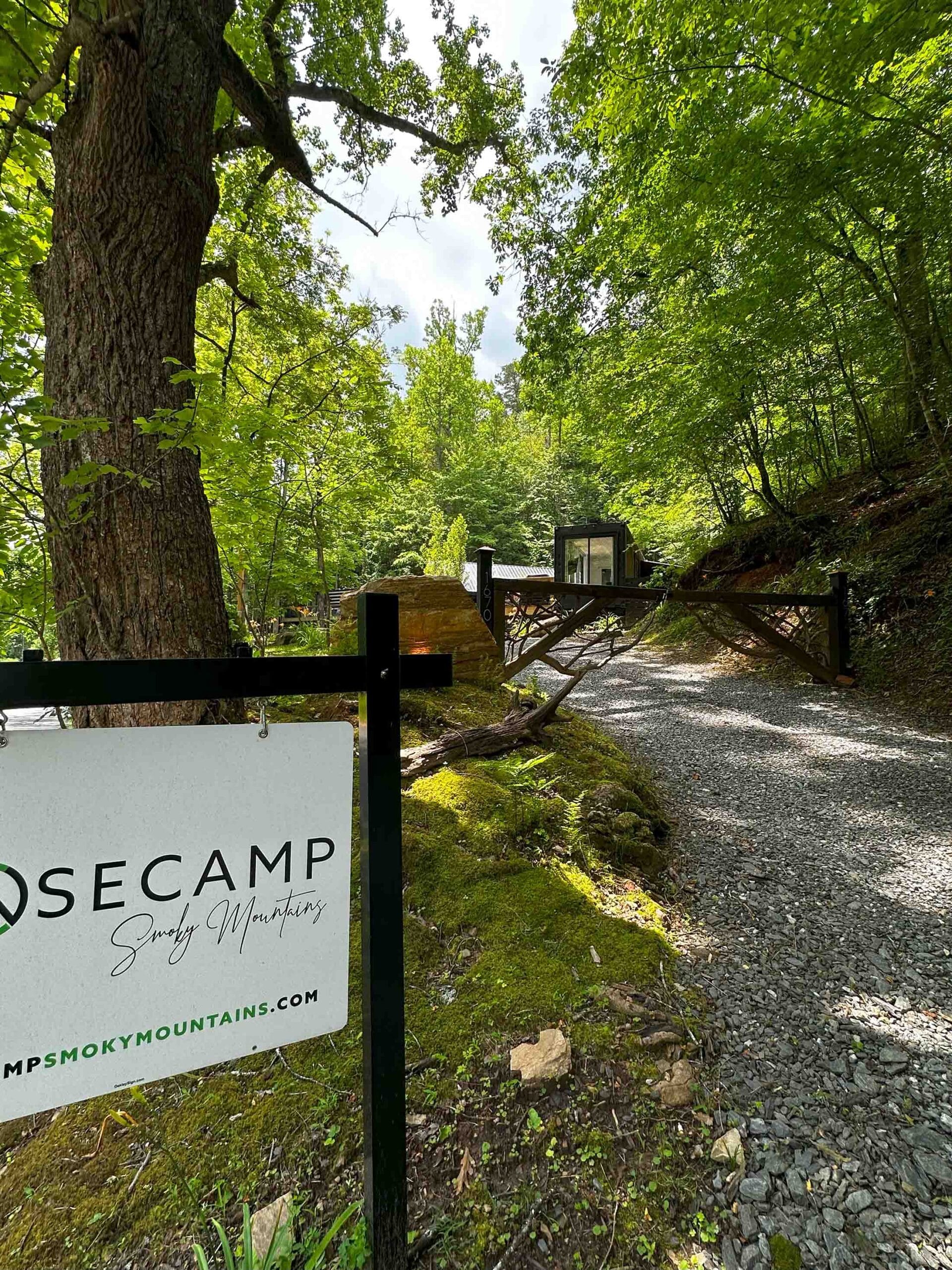 A gravel path leads to modern accommodations in a cabin nestled among green trees, with a sign in the foreground reading "OSE CAMP Smoky Mountains.