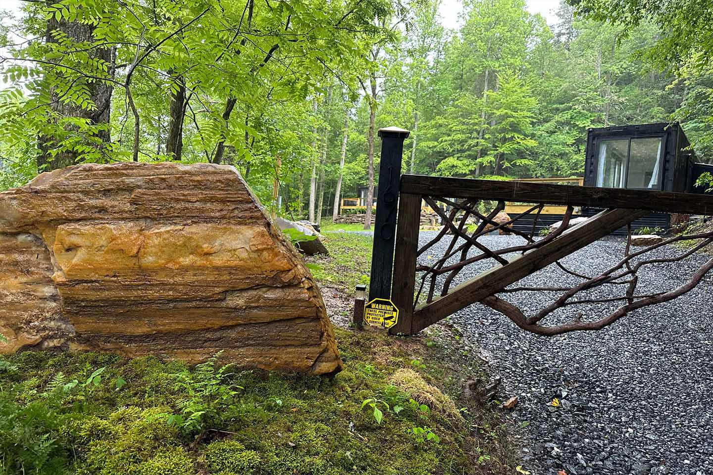 A wooden gate with a tree-branch design stands partially open on a gravel driveway, inviting new experiences amid green trees and a large mossy rock.