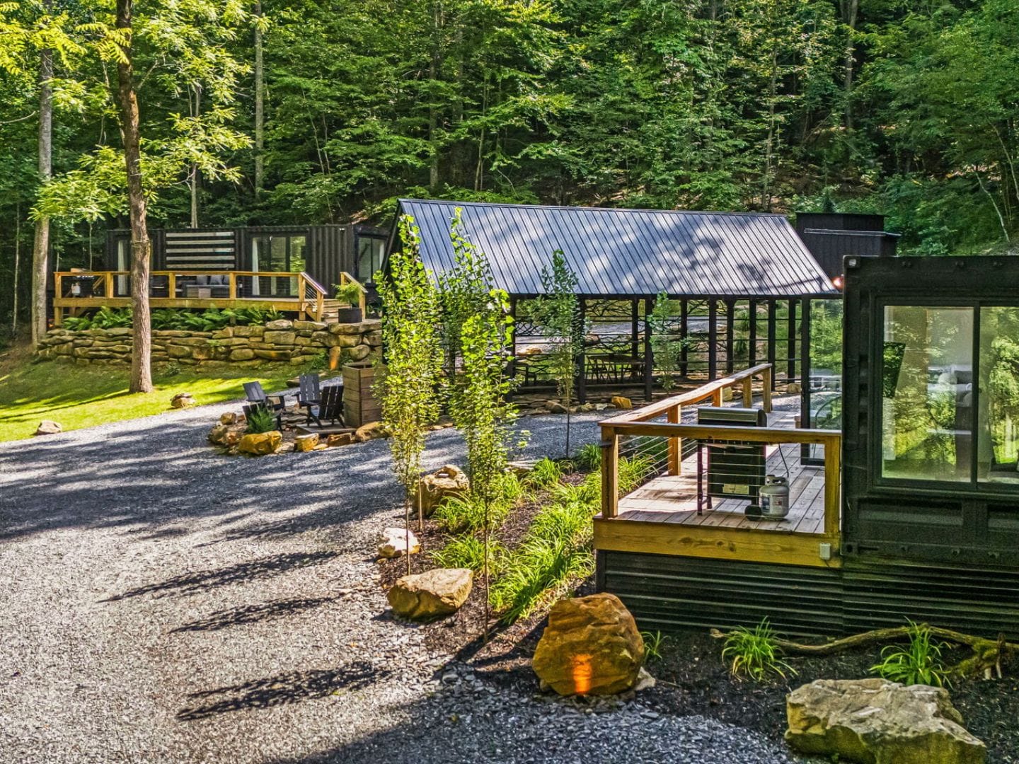 Outdoor area with a gravel path, wooden decks, a covered pavilion, seating around a fire pit, and surrounding trees in a forested setting.