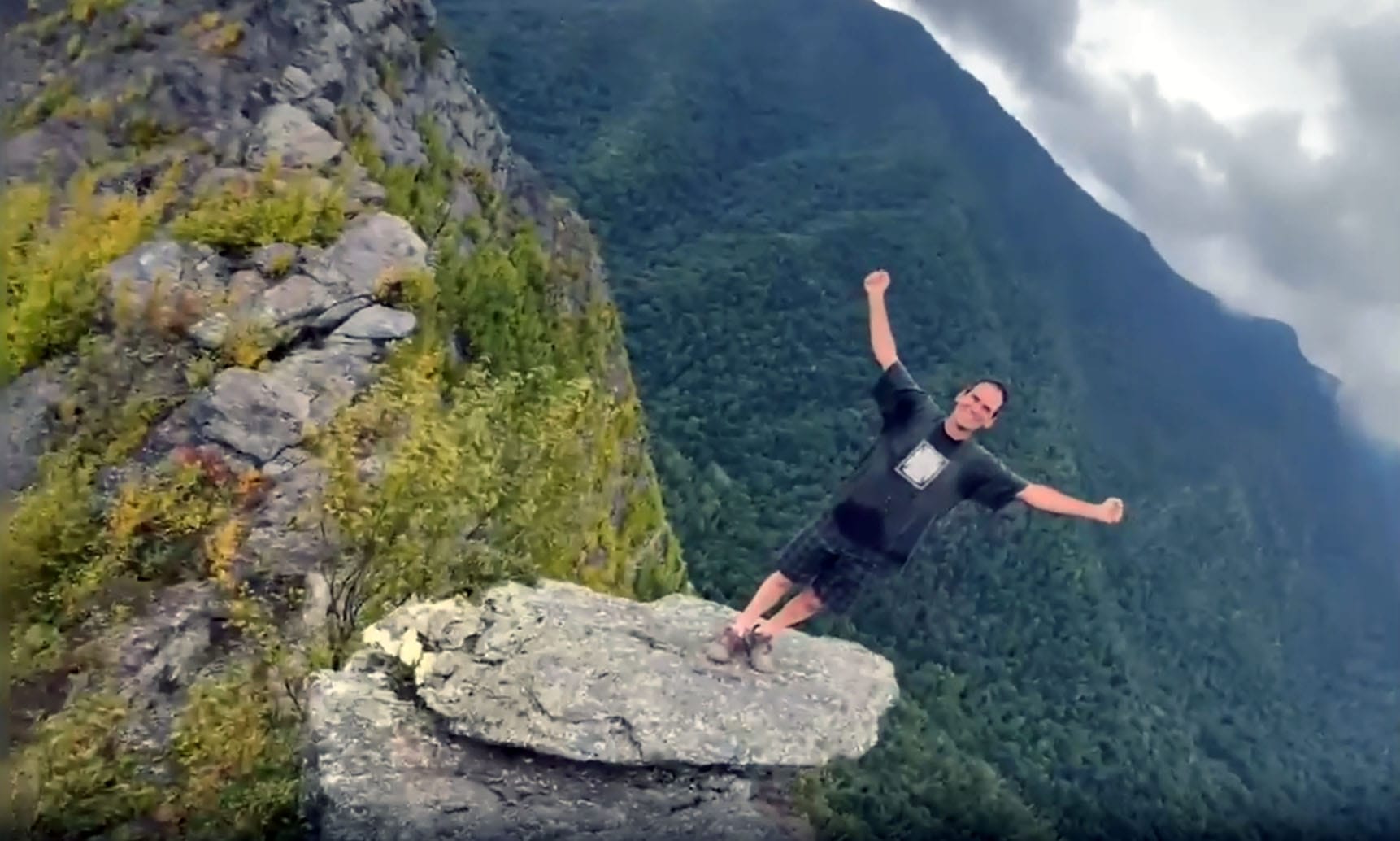 A person stands on the edge of a large rock outcrop with arms raised, surrounded by forested mountains and a cloudy sky—an inspiring scene from a nature retreat in the basecamp Smoky Mountains.