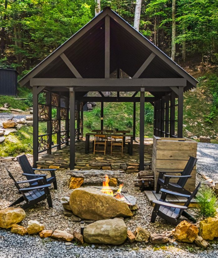 A gazebo in a wooded area with gravel paths leading to it, surrounded by chairs and a stone fire pit in the foreground.