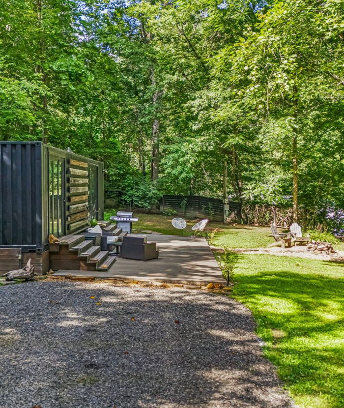 A secluded outdoor setting features a black shipping container home, a gravel driveway, several chairs, a fire pit, and surrounding tall green trees.