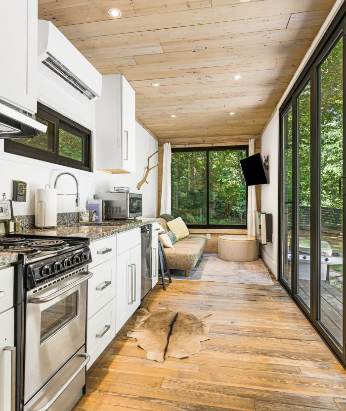 Modern tiny house interior with a compact kitchen on the left, featuring stainless steel appliances, white cabinets, and a wooden ceiling. Large glass doors and windows on the right overlook a green outdoor area.