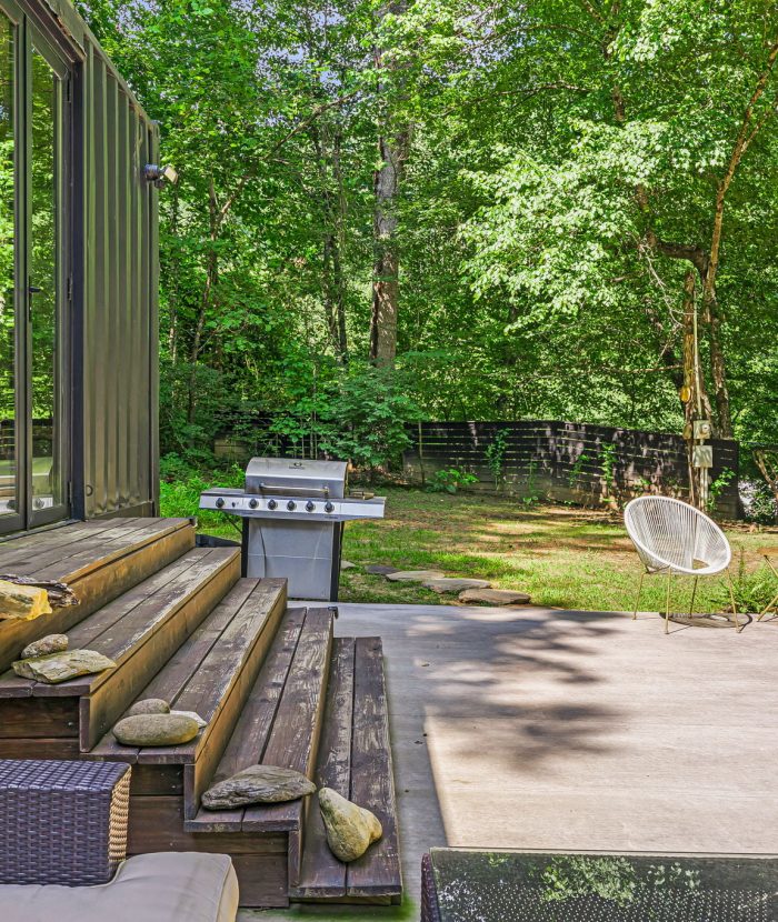 A patio with beige furniture, stone steps, a green vase, and a grill. Two white chairs are on a concrete area with surrounding greenery and trees.