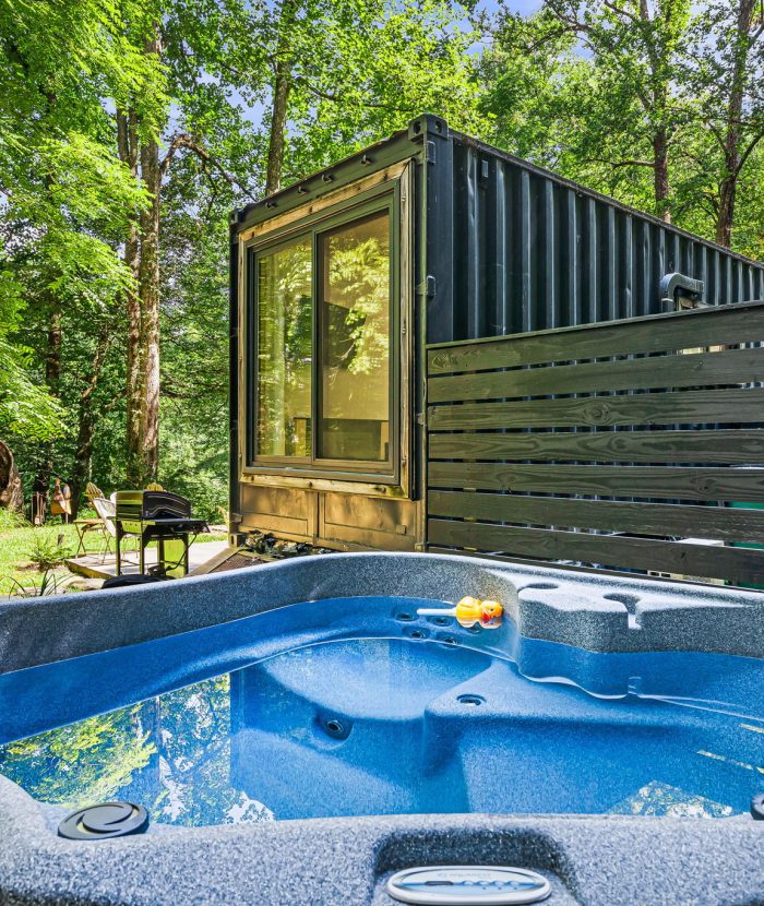 A hot tub is placed in front of a shipping container home, set in a forested area. The hot tub's blue water contrasts with the greenery of the surrounding trees.