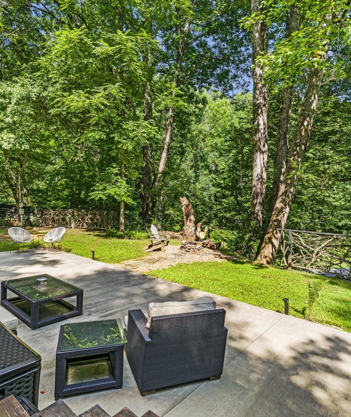 A spacious patio with outdoor furniture overlooks a lush green backyard bordered by a rustic wooden fence, surrounded by tall trees under a blue sky.