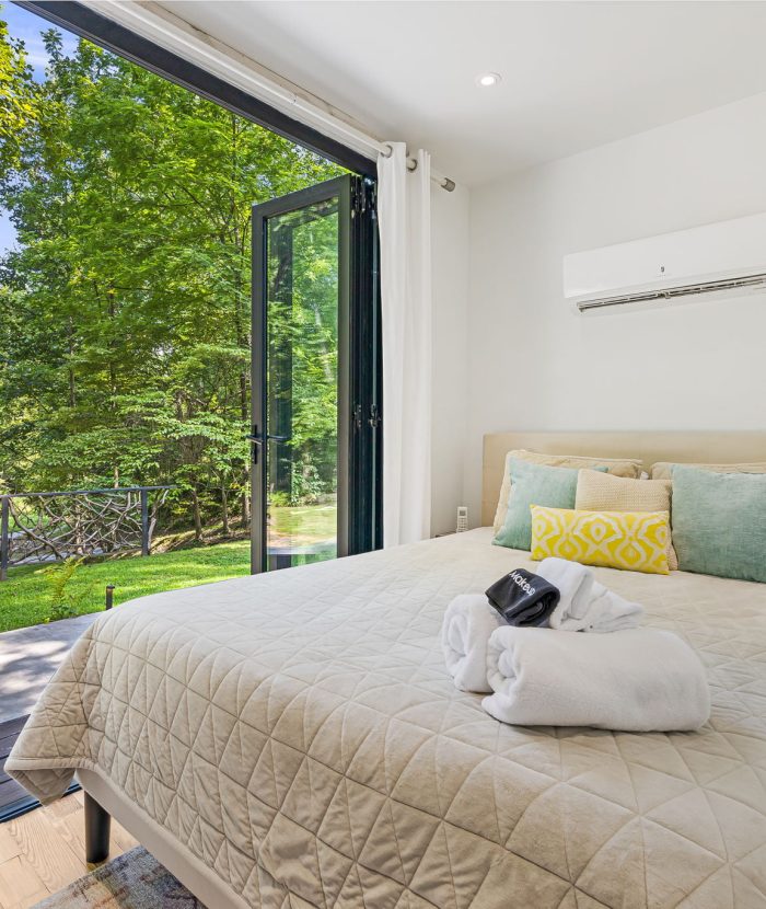 A bedroom with an open folding glass door leading to a deck and garden area. The room has a bed with pillows, a folded towel, and an air conditioning unit on the wall.