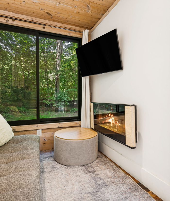 A cozy room features a grey couch, a wall-mounted TV, and a modern fireplace. Large windows allow a view of lush greenery outside. A round ottoman and neutral-toned decor complete the space.