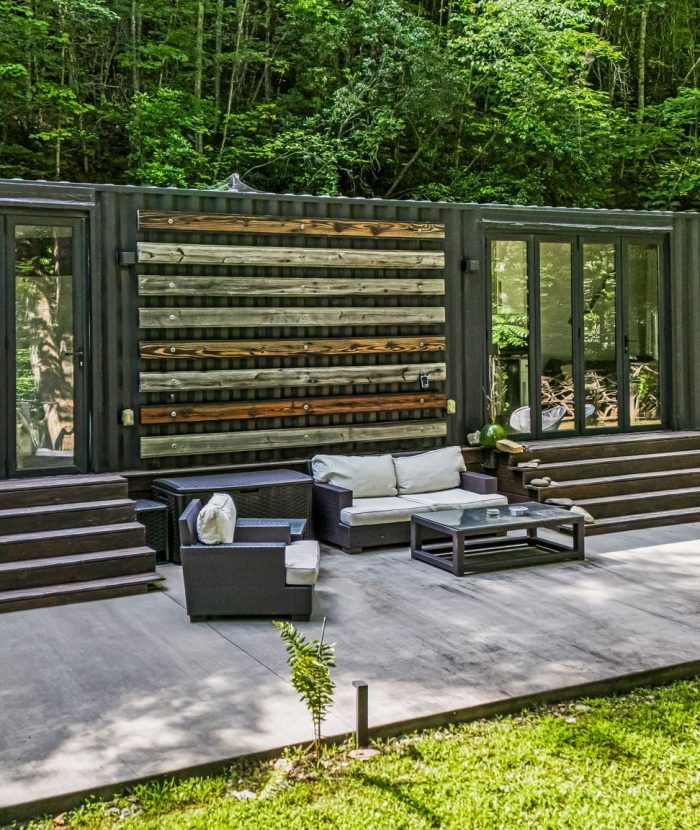 A modern container house with large windows, outdoor seating, and steps leading to a patio in a lush, wooded area.