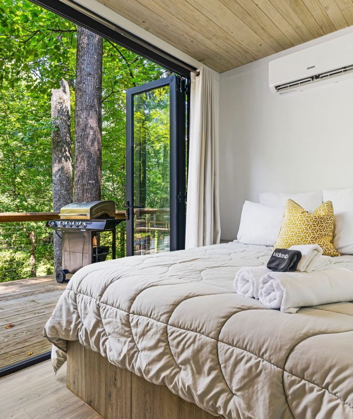 A cozy bedroom with a neatly made bed, adjacent to an open sliding glass door revealing a wooden deck with a barbecue grill and lush green trees outside.