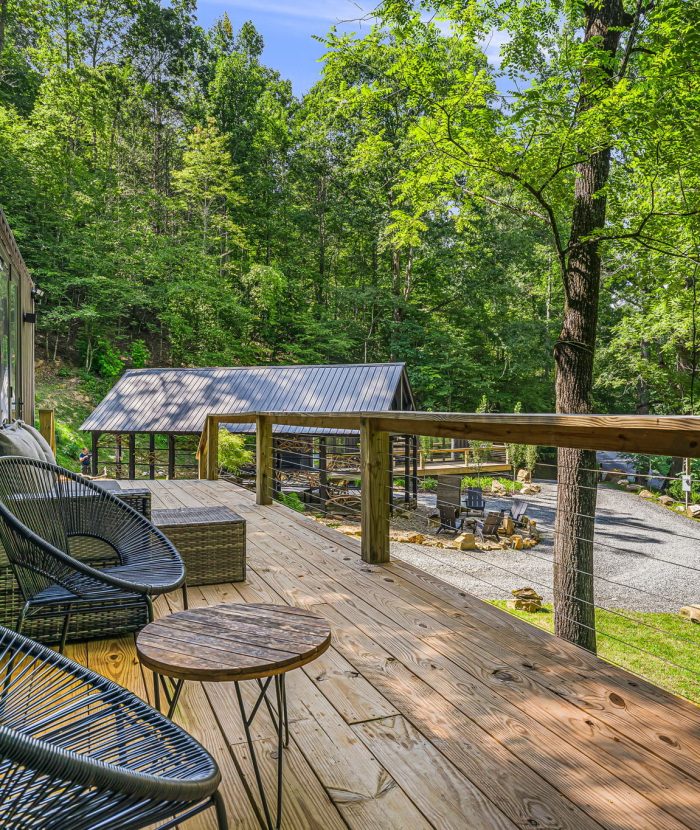 A wooden deck with outdoor seating overlooks a gravel pathway, lush green trees, and a metal-roofed structure amidst a forested area.