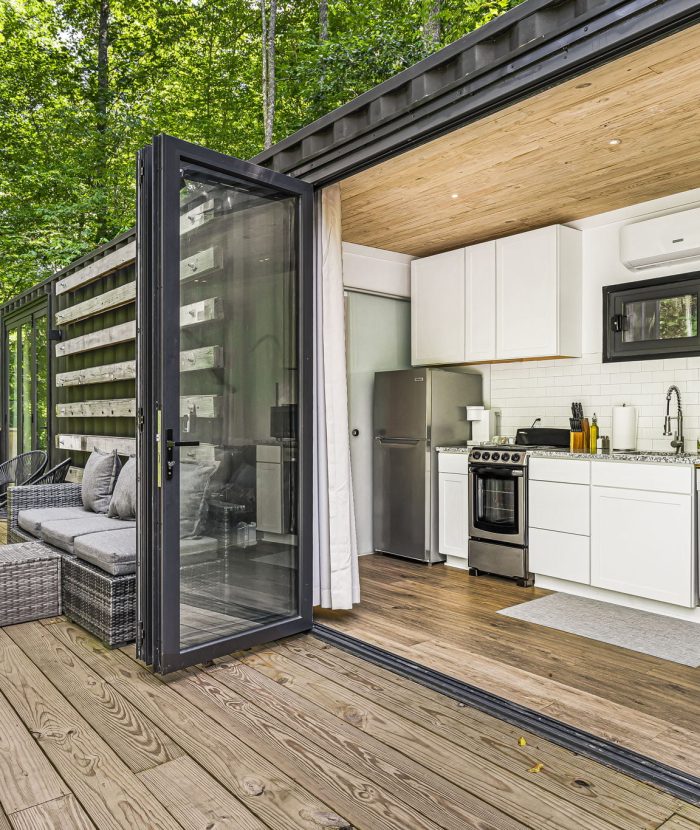 An open small house with a modern kitchen and a sitting area on a wooden deck surrounded by trees.