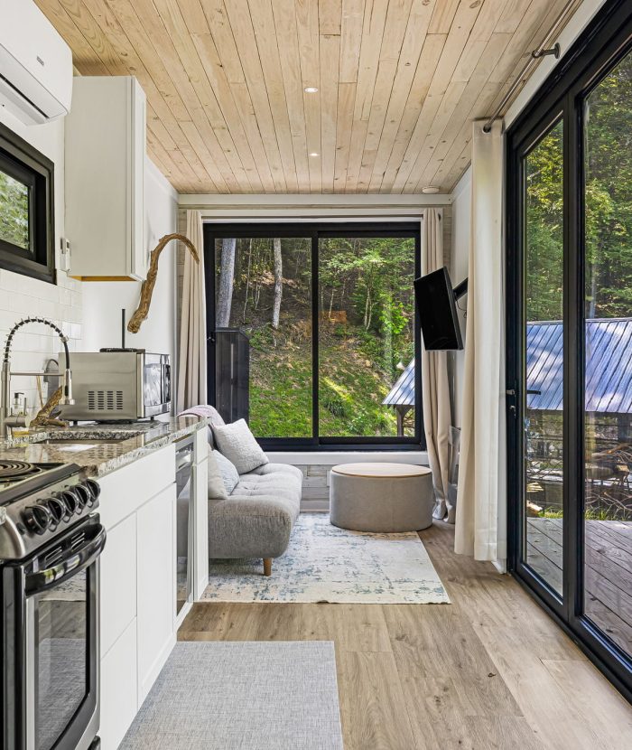 Modern tiny home interior with kitchen and living area featuring stainless steel appliances, a sofa, and a wall-mounted TV. Large sliding glass doors offer views of a deck and greenery outside.