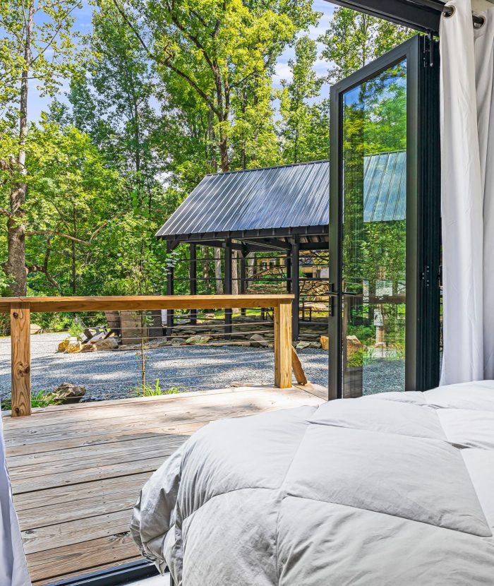 A bedroom with white bedding opens through glass doors to a wooden deck, overlooking a gazebo and a wooded area.