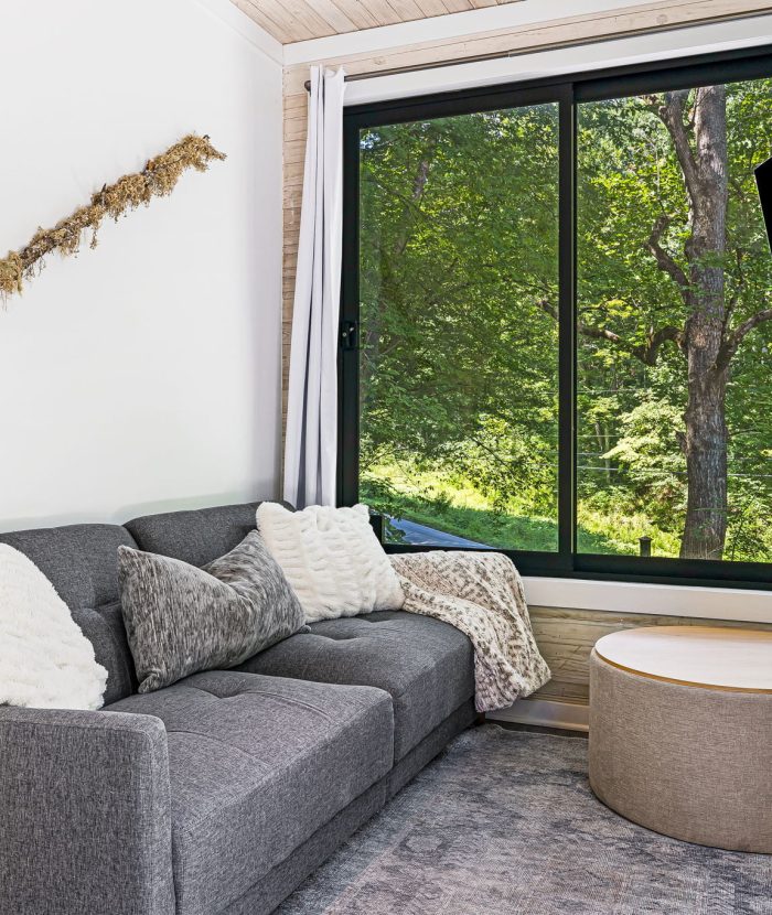 A cozy living room featuring a gray sofa with white and gray pillows, a round ottoman, a wall-mounted TV, a fireplace, wall art, and a large window with a view of a green forest outside.